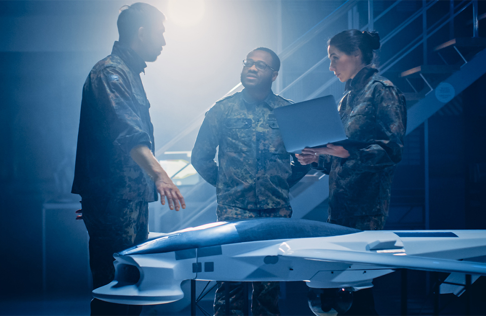 Army Aerospace Engineers Work On Unmanned Aerial Vehicle / Drone. Uniformed Aviation Experts Talk, Using Laptop. Industrial Facility with Aircraft for Performing Surveillance, Warfare Tactics, Attack.