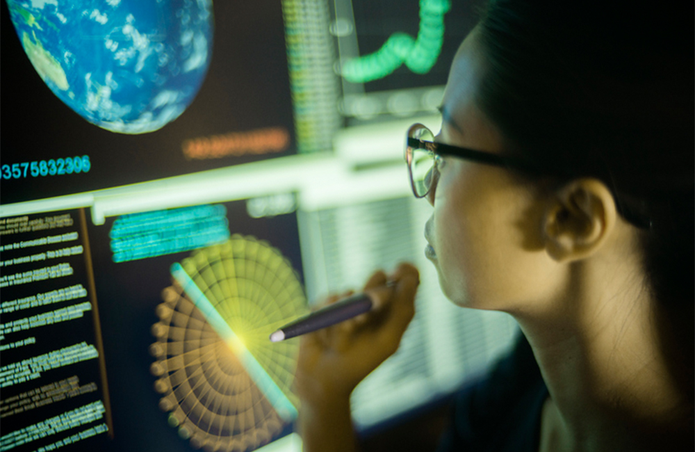 Stock photograph of a young woman, pointing at, and learning from, global data displayed on a large computer monitor.