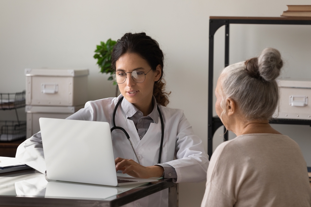 image of young female doctor meeting with elderly patient in office, listening to woman health problems complaints, typing on laptop, keeping electronic database on computer.