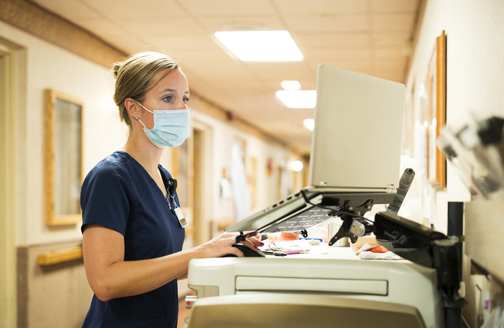 Registered nurse wearing a face mask documents medicine provided to a patient into a laptop computer at the nursing station at work, Indiana, USA