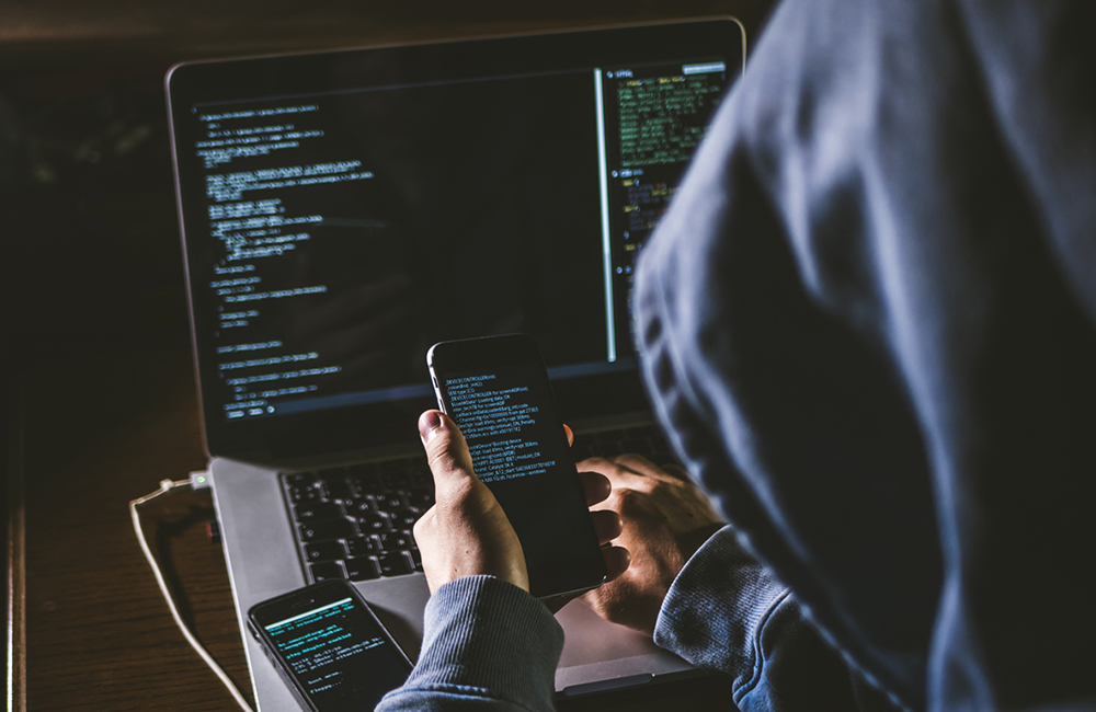 anonymous person in the hood sitting in front of computer working with laptop and mobile phone