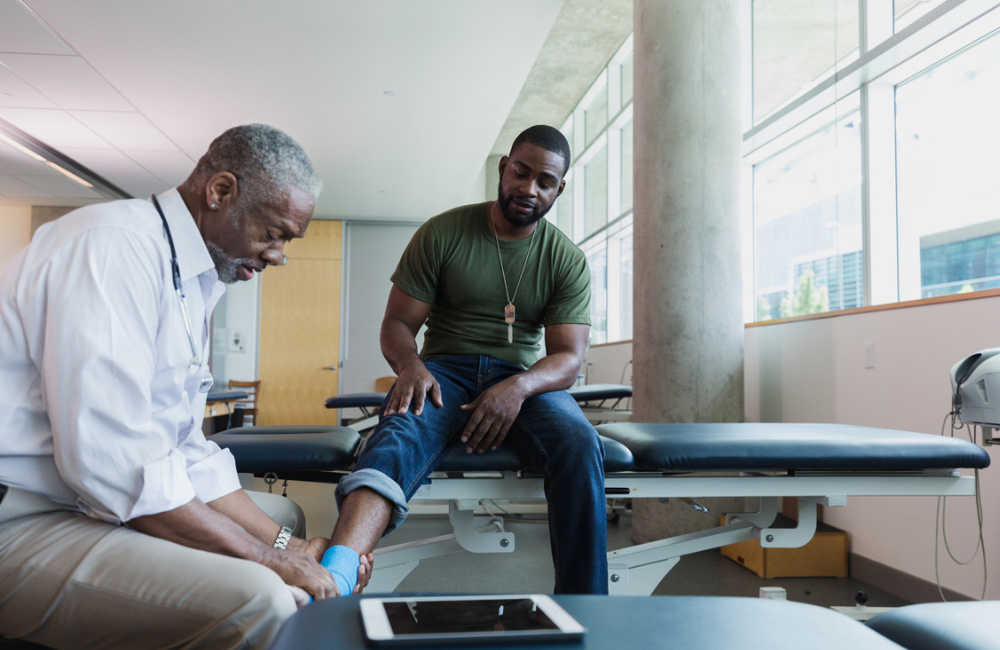 Serious senior doctor examines a mid adult male army soldier's injured ankle. The soldier's ankle is wrapped with sports tape.