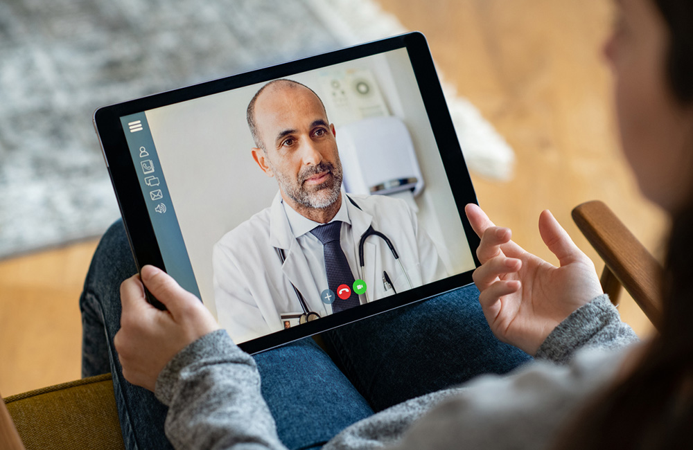 Back view of young woman making video call with her doctor while staying at home. Close up of patient sitting on armchair video conferencing with general practitioner on digital tablet. Sick girl in online consultation with a mature physician.